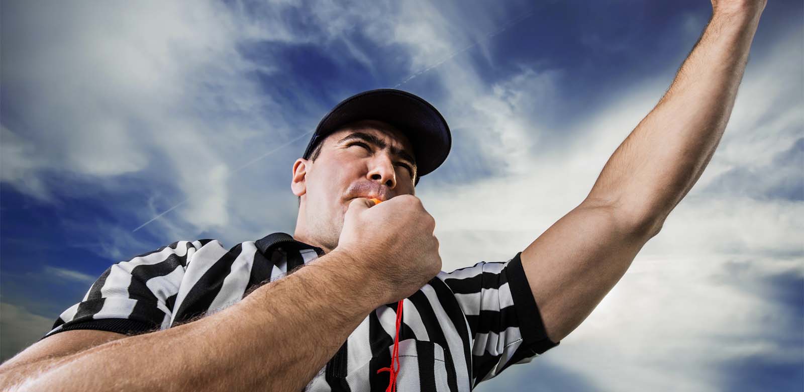 A referee standing outside with his arm raised and blowing a whistle.
